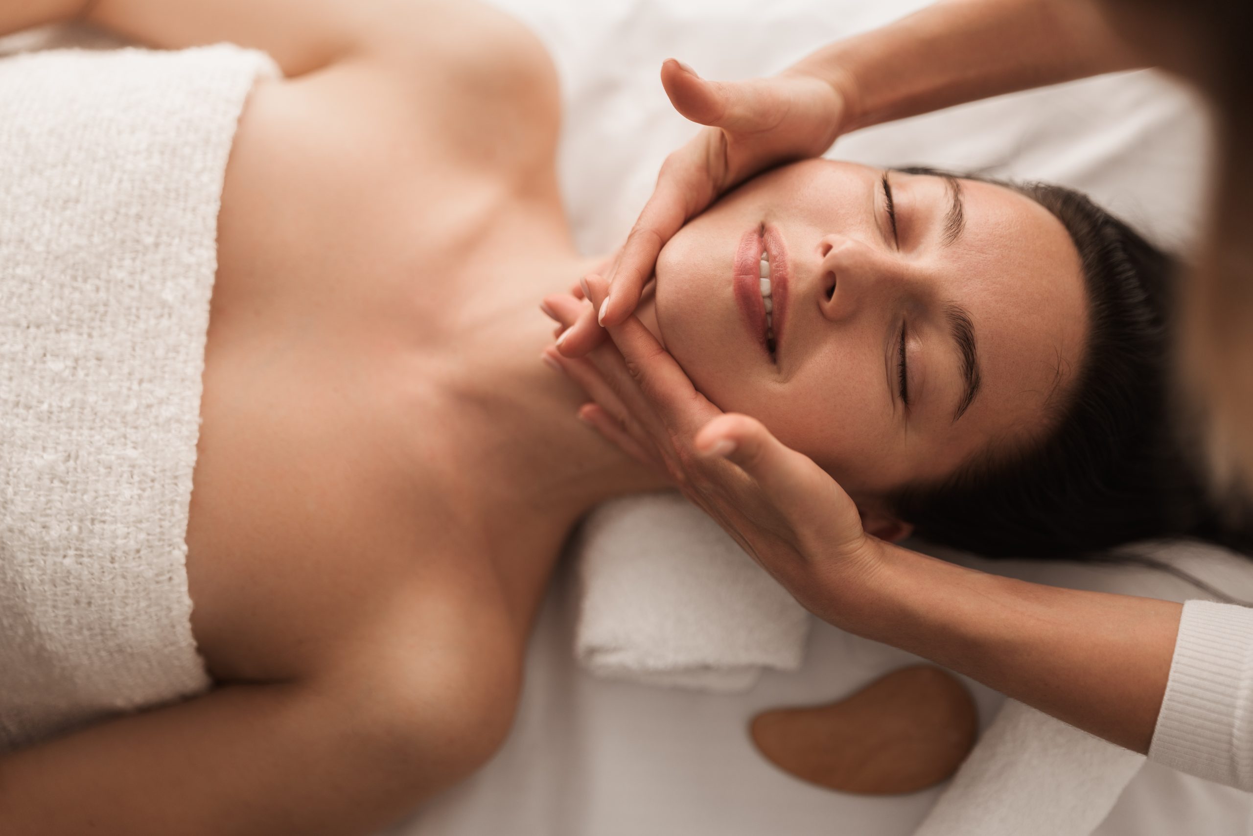 Top view of crop unrecognizable beautician doing face massage to delighted young female customer, lying on couch with closed eyes and smiling during spa treatment