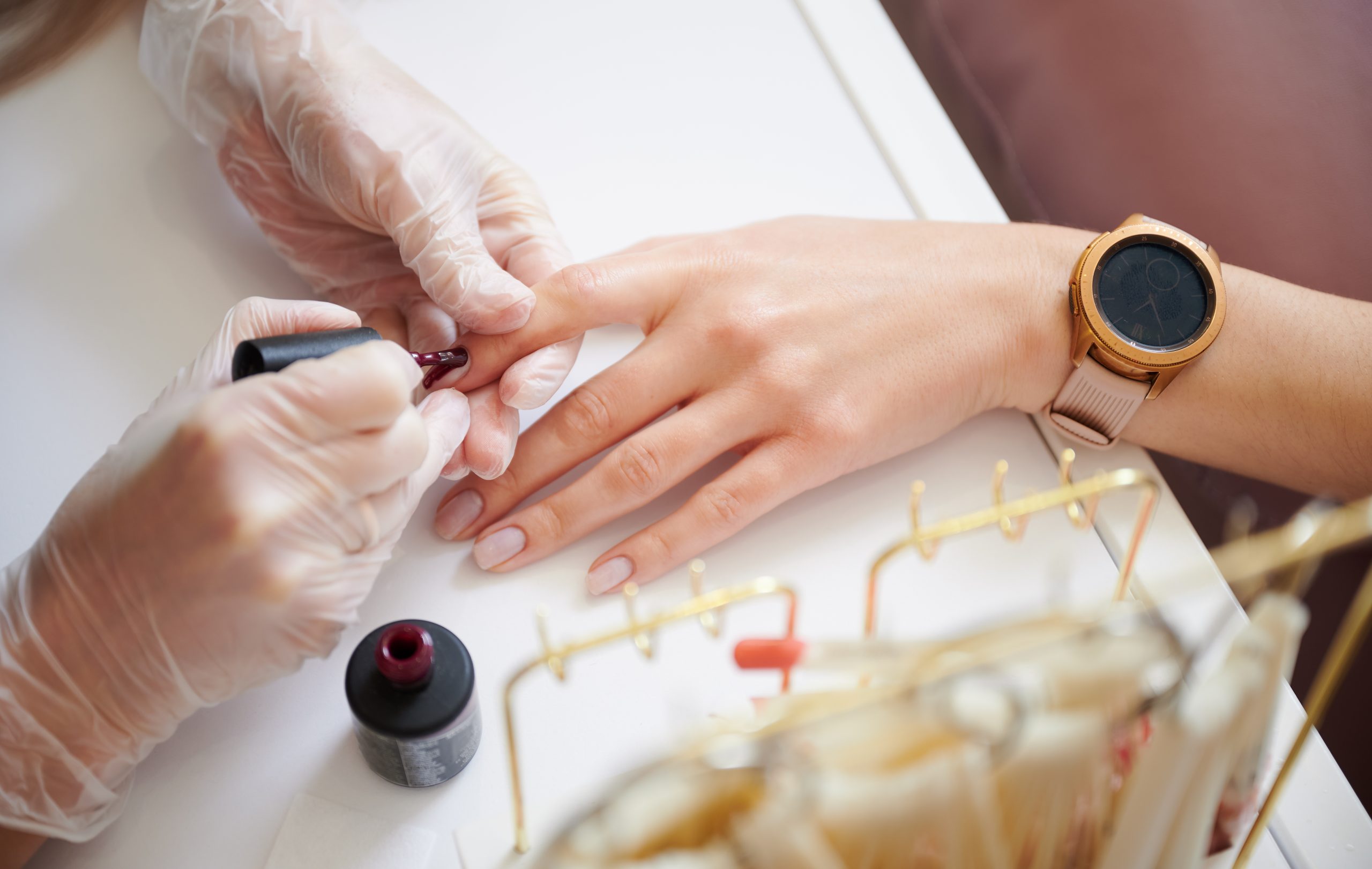 Professional manicurist doing manicure for woman client in beauty salon. Close up of manicure specialist hands in sterile gloves applying red nail polish on woman fingernail.